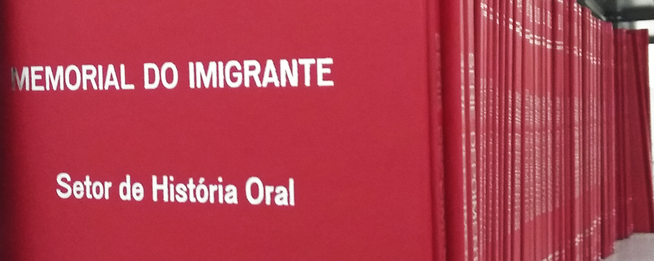 A imagem mostra, enfileirados, livros vermelhos, sendo que a capa do primeiro aparece. Está escrito 'Memorial do Imigrante' e 'Setor de História Oral'